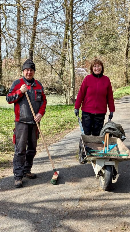 Zwei Personen auf einem Weg: einer mit Besen, die andere mit einer Schubkarre.